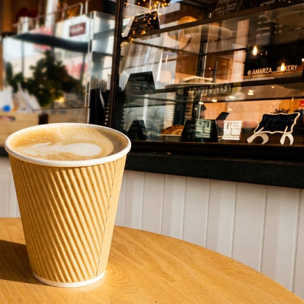 A latte in a paper cup sits on a wooden table with a bakery display case in the background.
