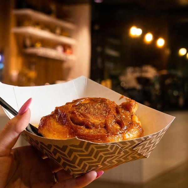 A hand holds a sticky bun in a paper tray with a fork, against a blurred restaurant background.