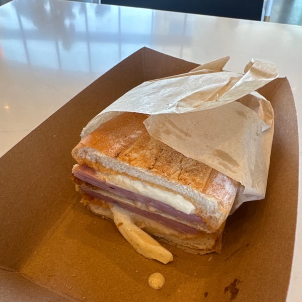 A pressed Cuban sandwich with ham, cheese, and mustard, partially wrapped in paper, in a cardboard tray.
