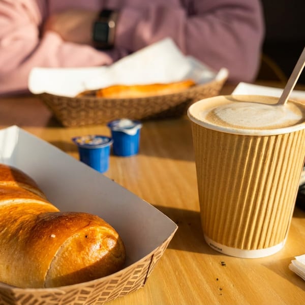 A table with a coffee cup, pastries in paper trays, and creamers, bathed in sunlight.